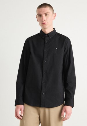 Black long-sleeve button-up shirt made of smooth fabric, featuring a classic collar, front placket, and embroidered logo on the chest.