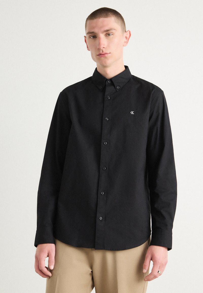 Black long-sleeve button-up shirt made of smooth fabric, featuring a classic collar, front placket, and embroidered logo on the chest.