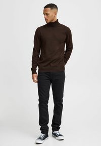 Maglione a collo alto marrone, texture a costine, vestibilità slim, abbinato a jeans neri e sneakers nere. Design semplice, senza motivi o decorazioni visibili.