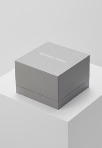 Scatola quadrata di cartone grigio con superficie texturizzata. "MICHAEL KORS" stampato in bianco sulla parte superiore. Si chiude con una cucitura orizzontale liscia.
