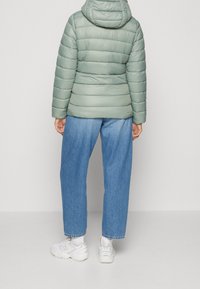 Lysegrøn dunjakke med vandret quiltning, fast hætte, kombineret med løse blå jeans og hvide sneakers. Blød tekstur, afslappet design.