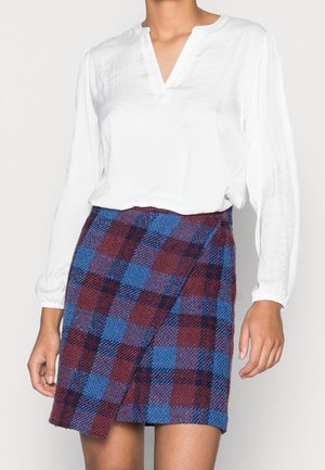 Femme portant un chemisier blanc à manches longues et une mini-jupe à carreaux bleue et bordeaux taille haute.