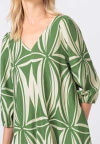 Green linen dress featuring a deep V-neck, voluminous puff sleeves, and a white abstract leaf pattern, with a textured fabric finish.