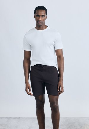 Άνδρας στέκεται φορώντας απλό λευκό κοντομάνικο πουκάμισο και μαύρο σορτς με κορδόνι, μπροστά σε ανοιχτό γκρι φόντο.