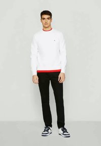 Balts garām piedurknēm krekls ar sarkanu apkakli, kombinēts ar melniem biksēm un tumši ziliem apaviem. Vienkāršs dizains, regulāra piegriezne, minimāla zīmola detaļa.