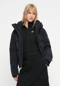 Blond kvinna som bär en svart vadderad jacka över en svart Adidas zip-up topp och svart kjol, ståendes mot en enkel vit bakgrund.