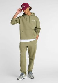 Olijfgroene hoodie met voorzak en logo, gecombineerd met bijpassende joggers. Aangevuld met een kastanjebruine pet en grijze sneakers. Eenvoudige, ontspannen pasvorm.