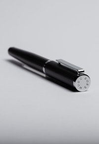 Bolígrafo negro con un barrel texturizado, clip de cromo y un extremo metálico con letras grabadas. Diseño elegante con una estética moderna.