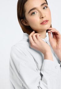 Jasnoszara koszula z kołnierzykiem o luźnym kroju, z detalami w postaci guzików i gładką teksturą tkaniny. Modelka delikatnie trzyma włosy jedną ręką.