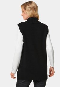 Schwarzer gestrickter ärmelloser Pullover mit hohem Kragen, ausgestattet mit Ripptextur und seitlichen Schlitzen, getragen über einem weißen Langarmshirt.