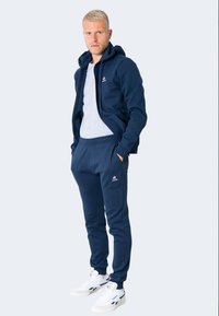 Felpa blu navy con zip, indossata sopra una t-shirt grigia, abbinata a joggers blu navy. Presenta dettagli con logo e sneakers bianche.