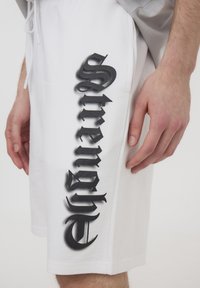 Shorts bianchi in cotone con stampa verticale in bold nero della parola "strength" sulla gamba sinistra, con una texture liscia e vita regolabile con cordoncino.