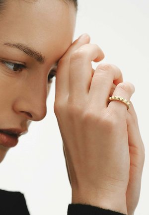 Primer plano de una persona con un anillo adornado con pirámides doradas en el dedo, descansando la mano cerca del rostro sobre un fondo liso.