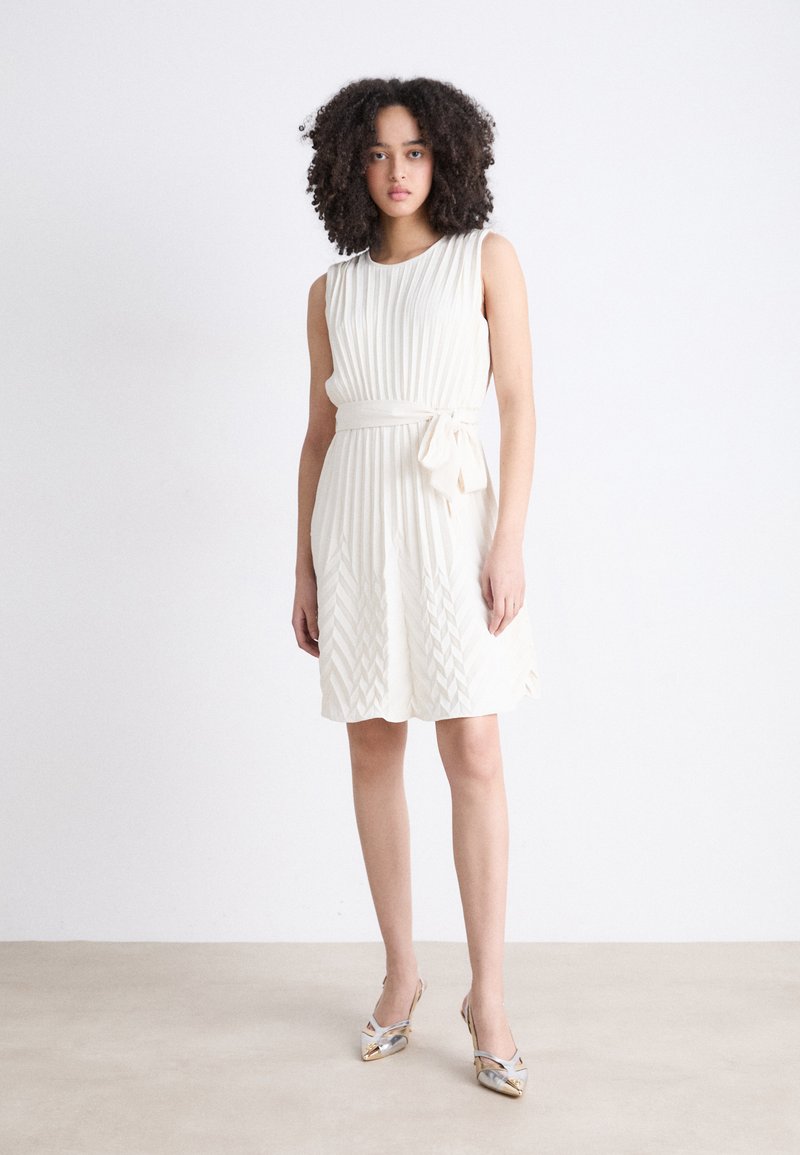 Cream-colored sleeveless dress with vertical pleats and a sash tied at the waist. Features a textured skirt with zigzag patterns.
