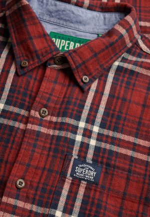 Chemise à carreaux rouge et bleu marine avec boutons marron, patch de la marque Superdry sur la poitrine et étiquette verte visible à l'intérieur du col.