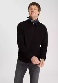 Czarny sweter z zamkiem błyskawicznym i długimi rękawami, wykonany z lekkiego materiału, z prążkowanymi mankietami i subtelną naszywką z logo. W zestawieniu z ciemnoszarymi dżinsami i niebieską chustą.