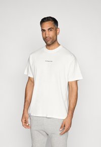 T-shirt branca de algodão com mangas curtas e decote redondo. Apresenta um pequeno detalhe em texto azul-escuro no peito. Textura suave, corte relaxado.