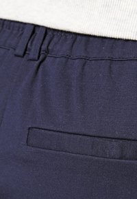 Gros plan sur un pantalon bleu marine avec une taille élastique et une poche arrière horizontale, porté avec un haut de couleur claire.