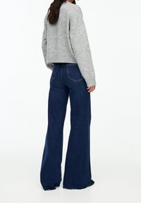 Suéter corto de punto gris con mangas anchas, combinado con jeans acampanados oscuros de talle alto que tienen un dobladillo sin terminar y bolsillos traseros.
