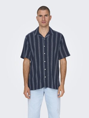 Kurzarmhemd mit Kragen in Marineblau mit weißen vertikalen Streifen, mit durchgehender Knopfleiste und strukturiertem Stoff. Trägt dazu eine helle Jeans in Blau.