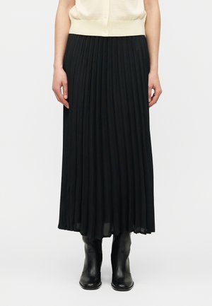 Vrouw die een zwarte geplooide midi-rok draagt, gecombineerd met zwarte leren laarzen en een crèmekleurige mouwloze top, staand tegen een effen achtergrond.