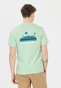 Vaaleanvihreä t-paita, joka on valmistettu pehmeästä puuvillasta. Paidassa on selässä graafinen design, jossa on vuoria, raitoja ja palmuja sinisenä ja valkoisena.