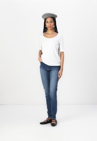 Hvid kortærmet top med rund halsudskæring, blå slim-fit jeans, grå beret og sorte flade sko med stropper, stående på en hvid baggrund.