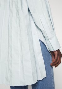 Boční pohled na osobu v světle modré pruhované dlouhé košili se bočními knoflíky a světle modrých džínách, ruka uvolněně položená vedle těla.