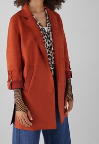 Blazer de ante color óxido con mangas enrolladas, dos bolsillos frontales y un corte relajado, superpuesto sobre una camisa de estampado de leopardo y mangas de red.