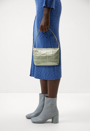 Sac à main matelassé vert clair avec une courte anse, tenu par une personne portant une robe bleue texturée, assortie de bottines grises à la cheville.