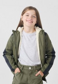 Giacca verde oliva con fodera crema soffice, maniche lunghe con dettagli neri, chiusura frontale con cerniera, indossata sopra una maglia grigia e pantaloni verdi.