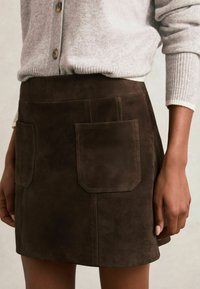 Barna velúr szoknya, két nagy foltvarrott zsebbel, függőleges varrásokkal és testhezálló fazonnal, világosszürke kötött pulóverrel párosítva.