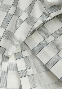 Λευκό ύφασμα με σχέδιο πλέγματος από λεπτές μαύρες γραμμές και μικρές μαύρες διακεκομμένες λεπτομέρειες, που δείχνει πτυχώσεις και υφή.