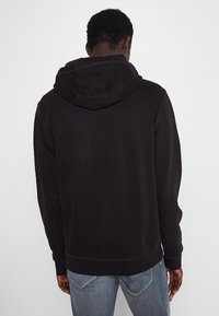 Mężczyzna w czarnej bluzie z kapturem i niebieskich dżinsach, stojący tyłem na jednolitym jasnym tle.