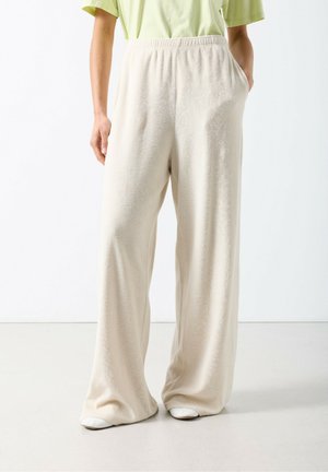 Pantaloni beige a gamba larga con finitura morbida e testurizzata, vita elasticizzata e tasche laterali, abbinati a una maglietta verde a maniche corte.