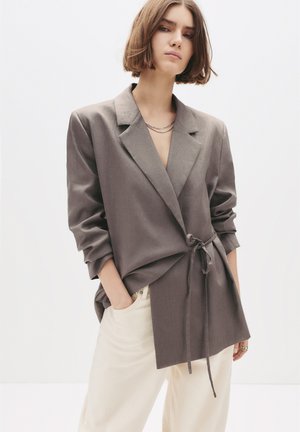 Femme portant un blazer gris oversize noué à la taille, un pantalon beige clair, avec des cheveux bruns courts et un collier en chaîne simple.