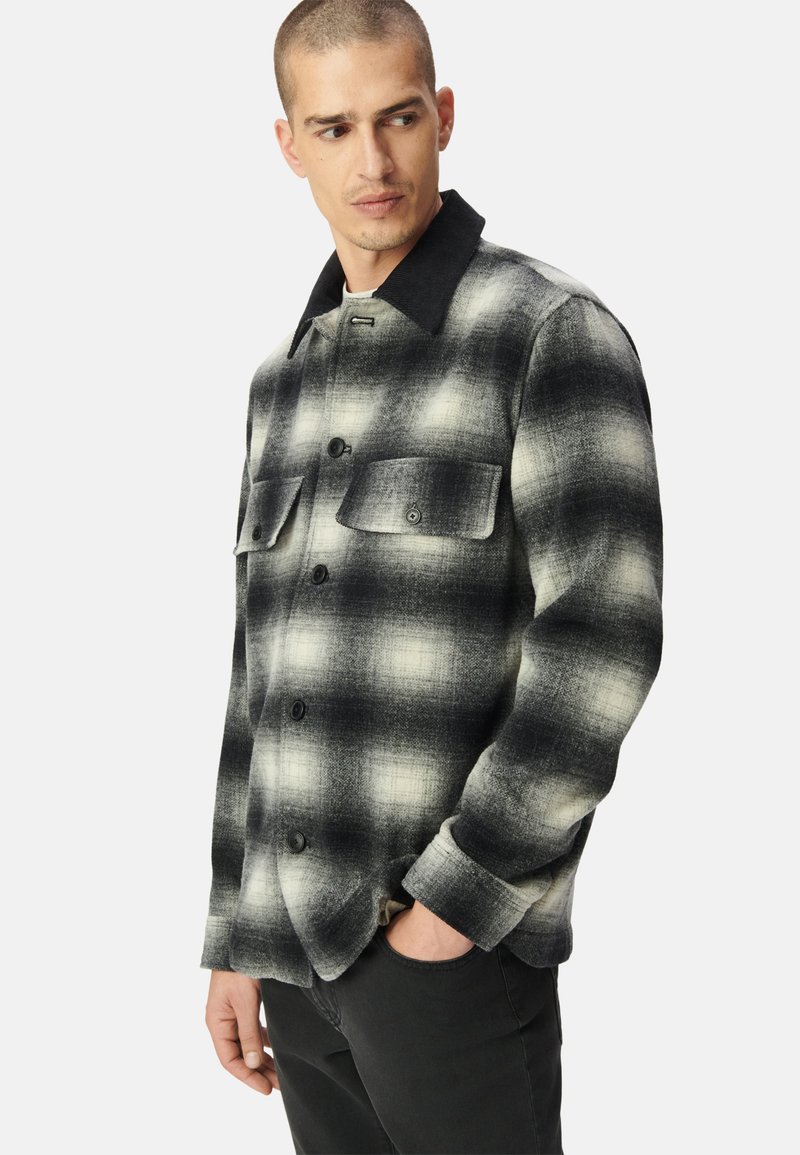 Camicia in flanella a quadri bianca e nera con colletto, due tasche sul petto e bottoni neri. Materiale strutturato con una finitura pattern.