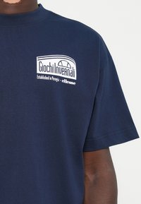 Marineblå t-shirt lavet af bomuld med et hvidt grafisk logo, der viser "Giochi Invernali" og brandnavnet "ellesse" nedenfor. Kortærmet.