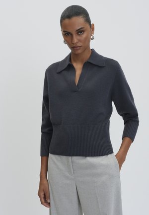 Vrouw met donker haar in een lage knot, gekleed in een donkergrijze gebreide trui met kraag en lichtgrijze broek, staand met één hand in de zak.