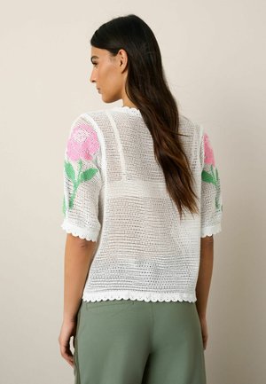 Žena s dlouhými tmavými vlasy, oblečená v bílém síťovaném svetru s růžovými květinovými vzory na rukávech a zelených kalhotách, viděná zezadu.