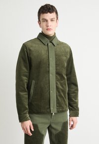 Green corduroy jacket with a collar, snap buttons, and side pockets. The fabric has a textured ribbed pattern, paired with a matching colored turtleneck.