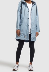 Giacca impermeabile azzurro chiaro con cappuccio, cerniera frontale, bottoni a pressione e tasche laterali. Abbinata a leggings neri e scarpe da ginnastica.