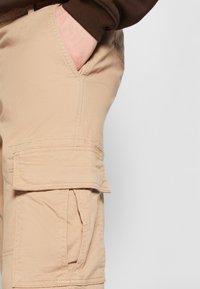 Pantalon cargo beige avec une grande poche latérale, des coutures courbes et une texture lisse. La poche est dotée d'un rabat et de coutures visibles.