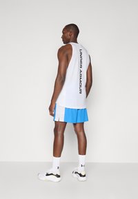 Valkoinen hihaton toppi, jossa on musta "UNDER ARMOUR" -teksti ja siniset urheilushortsit. Sisältää rennon leikkauksen ja sivuleikkaukset liikkumista varten.