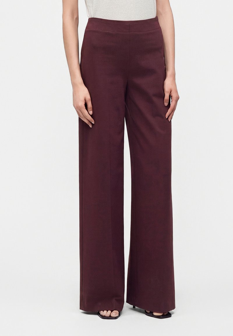 Personne portant un pantalon bordeaux taille haute à jambes larges, associé à des talons noirs ouverts et un haut sans manches de couleur claire.