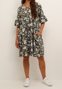 Olivegrünes Kleid mit weißem Blumenmuster, locker geschnitten, kurze Rüschensäume und seitliche Taschen. Kombiniert mit weißen Sneakers.