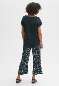 Losse, donkergroene top met opgerolde mouwen, gecombineerd met brede zwarte broek met een mintgroen abstract patroon. Zwarte sandalen.