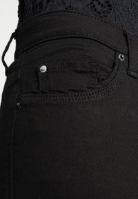 Jeans de mezclilla negra que presentan un primer plano de un bolsillo frontal con detalles de herrajes plateados y costuras. La tela parece suave y ligeramente texturizada.