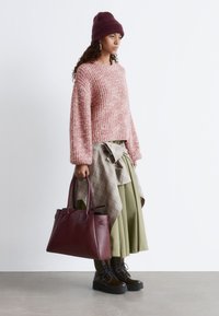 Rosa gestrickter Pullover mit weiten Ärmeln, karierte Bluse in Schichten um die Taille gebunden über einem grünen Rock, burgunderfarbene Handtasche und schwarze Combat-Stiefel.