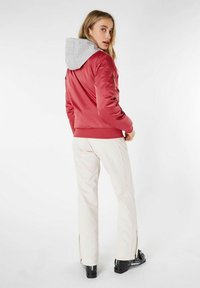 Chaqueta acolchada roja con una sudadera gris, puños y dobladillo acanalados, y bolsillos laterales con cremallera, combinada con pantalones blancos y calzado negro.